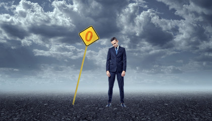 Upset businessman standing on rocky ground in front of yellow road sign with zero