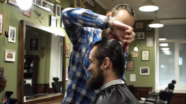 Long hair barber is doing a haircut to young bearded man in barbershop. 4K
