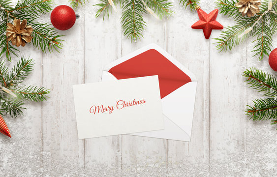 Merry Christmas greeting on white paper. Envelope beside. Christmas tree with decorations beside. White wooden table in background.