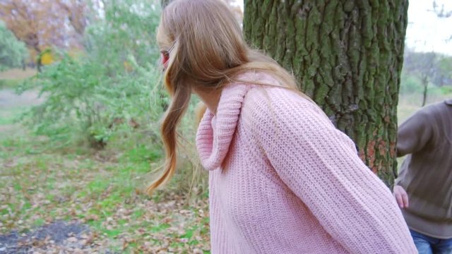 Attractive blonde girl hiding in the forest and leaning against the old tree, playing