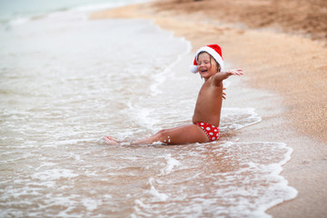 tanned Caucasian kid girl in santa hat on  beach with yellow san