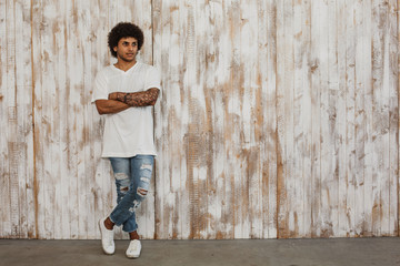 Beautiful young dark-skinned man with curly hair stands, against retro wooden wall. Modern youth style clothes.