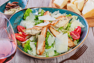 Salad with chicken breast, parmesan cheese, croutons, tomatoes, mixed greens, lettuce and glass of wine on light wooden background. Ingredients on table