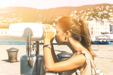 Young woman tourist looking through binoculars at horizon enjoying the beautiful sunny day - Outdoor portrait of stunning girl in a summer day