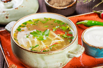 Vegetable soup, broth with noodles, herbs, parsley and vegetables in bowl with sour cream, spice, pepper, dried thyme and bread on dark wooden background. Ingredients on table