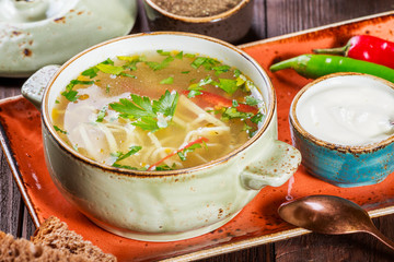 Vegetable soup, broth with noodles, herbs, parsley and vegetables in bowl with sour cream, spice, pepper, dried thyme and bread on dark wooden background. Ingredients on table