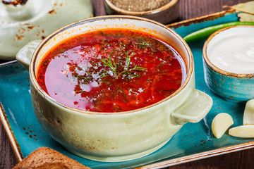 Ukrainian and Russian traditional beetroot soup - borscht in clay pot with sour cream, spice, garlic, pepper, dried herbs and bread on dark wooden background, healthy food. Ingredients on table.