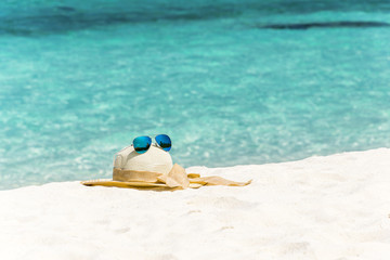 Fototapeta na wymiar White female trendy hat with blue aviator sunglasses lying on white sand beach in front of turquoise ocean. 