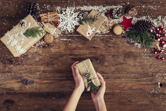 Overhead view of a Girl's hands holding a wrapped Christmas gift with assorted gifts and decorations on a wooden table