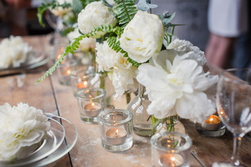 Nice rustic wedding table decorated with beautiful white David Austin roses and candles in glass holders. Rare romantic flowers on plates. Loft style wedding, blurred background.