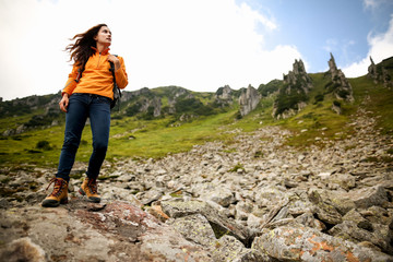 Naklejka na ściany i meble Young woman traveler with backpack relaxing outdoor with rocky mountains on background. Lifestyle hiking concept. Backpacker hiking at mountain peak