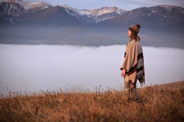 Young woman hiking at mountain peak above clouds and fog Hiker girl wrapping in warm poncho outdoor. Misty mountain. Young girl over the clouds in the valley looking at calm autumn sunrise