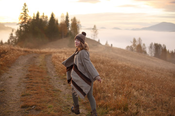 Naklejka na ściany i meble young woman hiking at mountain peak above clouds and fog Hiker girl wrapping in warm poncho outdoor.Early morning.Misty mountain.Young woman in a dress running to the sun in the mountains. Boho style