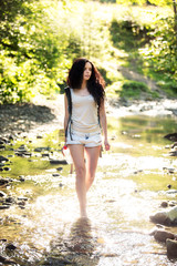 Outdoor shot of attractive young woman with backpack standing in a mountain stream. Female hiker in creek water.Portrait of young pretty brunette woman holding bottle of water.