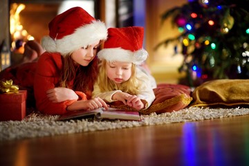 Fototapeta na wymiar Two cute little sisters reading a story book together under a Christmas tree