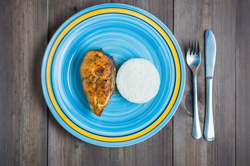 blue turquoise round plate with red fried fish, rice, fork and knife on dark wooden background view from the top
