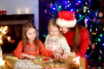 Fototapeta premium Two adorable little sisters and their mother baking Christmas cookies by a fireplace