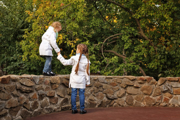 Fototapeta premium Cute little sisters in the autumn park. The older girl helping younger go on the stone fence. Family time. Sport games.