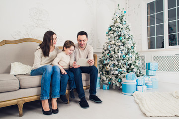 Obraz premium Cheerful family sitting in the living room having fun with the digital tablet that Santa Claus brought her, behind the decorated christmas tree