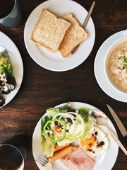 Overhead view of Hotel Breakfast - Soft boiled rice with fried eggs, bacon, sausages, ham, fresh salad, toast and hot tea on top a wooden table 