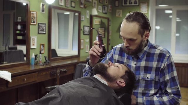 Attractive bearded man getting his beard shaved by modern barber in barber shop