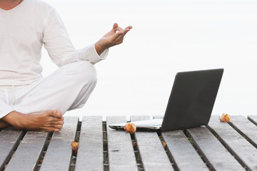 Man in white clothes meditating yoga with laptop on wooden pier