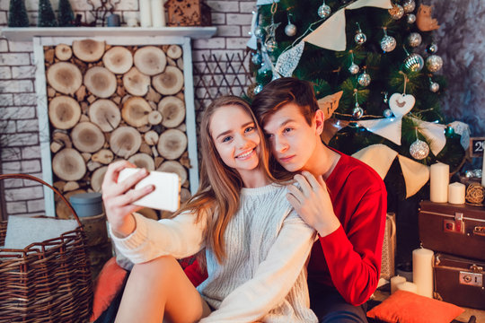 Young couple in love sitting near christmas tree having fun and make selfie. New Year