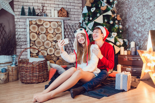 Young couple in love sitting near christmas tree in funny hats having fun and make selfie. New Year