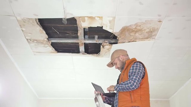 young man makes an emergency inspection of ceiling and use laptop PC. public utilities. Ceiling panels damaged huge hole in roof from rainwater leakage.Water damaged ceiling , Insurance agent.