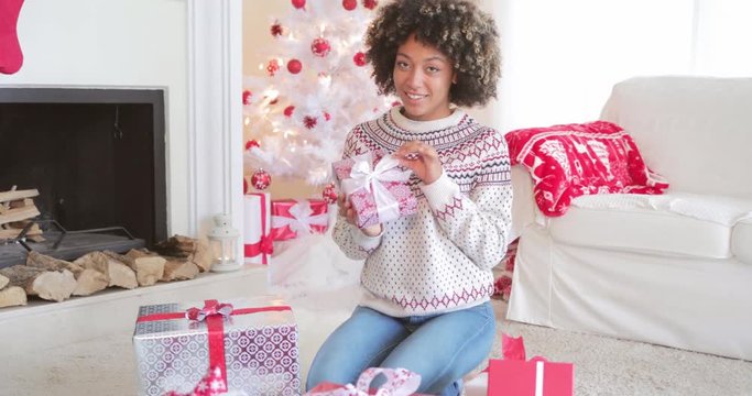 Pretty happy young woman checking her Christmas gifts on the floor in her living room holding up a decorative box tied with a bow with friendly smile.