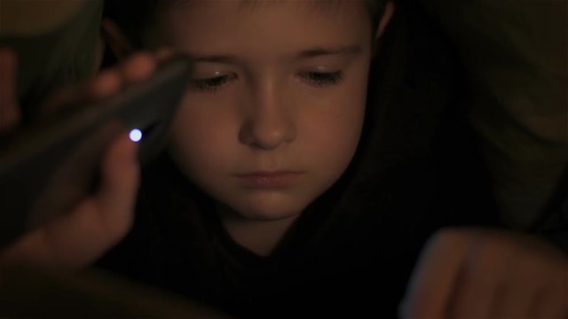 Six year old boy read book under covers at night and illuminates flashlight. Close up. Little boy lying under covers at night. Teenager under covers playing on tablet and reading book