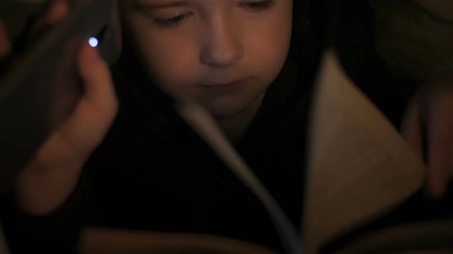 Boy reading book before going to sleep under covers. Close up. Little boy lying under covers at night. Teenager under covers playing on tablet and reading book