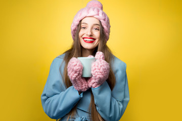 Woman in colorful winter clothes holding a cup of hot drink standing on the yellow background. Happy winter concept