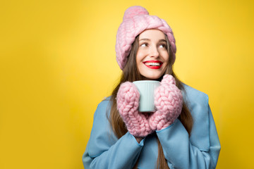 Woman in colorful winter clothes holding a cup of hot drink standing on the yellow background. Happy winter concept