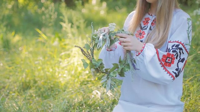 Slavic woman in an embroidered shirt patterns , weaves a wreath of wild flowers sitting by the fire .