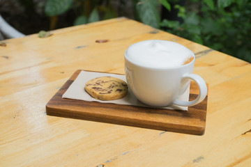 hot coffee in white cup white foam chocolate chip cookie biscuit with silver spoon on wooden table at coffee time sunset / hot coffee cookie