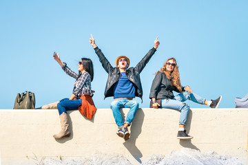 Exultant friends with man hands up and happy woman freezing the moment taking selfie - Students joy and victory gesture sitting together under blue sky background - Concept of people good emotions