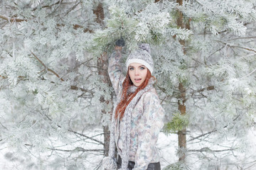 Fototapeta na wymiar beautiful cheerful happy girl with red hair in a warm hat and scarf playing and fooling around in the snow in the winter forest