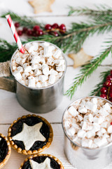 Hot cacao in metal cup covered with melting marshmallows, tarts with jam and berries on white wooden table