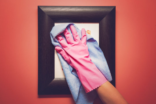 Hand in pink protective glove wiping picture or photo frame on the wall from dust with rag. Early spring cleaning or regular clean up. Maid cleans house.