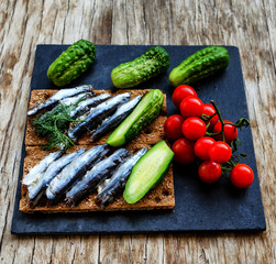 Anchovy with fresh cucumbers and cherry tomatoes on the black rectangular plate on the vintage wooden table, traditionally accompanied with vodka