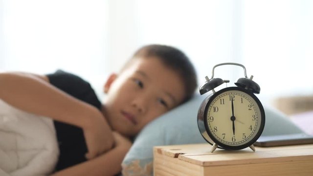 Selective focus at vintage alarm clock show at six o clock and out focus young asian boy sleeping and wake up on the bed.