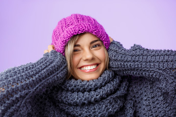 Young beautiful fair-haired girl in knited hat and sweater smiling looking at camera over violet background. 