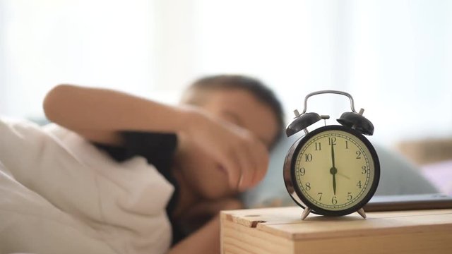 Selective focus at vintage alarm clock show at six o clock and out focus young asian boy sleeping and wake up to play smart phone on bed.
