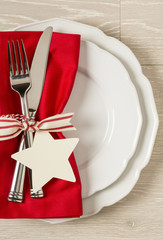 Festive Christmas table setting place setting with white china plates, red cloth napkin and silverware on rustic wood background. Vertical with empty blank space on right side.