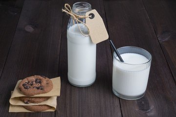 bottle of milk with carton label tag with one transparent glass, black straw and few brown cookies on dark wooden background