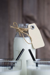 closeup shot bottle of milk with carton label tag with two transparent glasses, black straw on dark wooden background