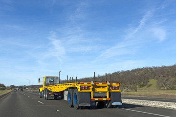 Rear and side view of yellow flatbed semi on highway under blue sky. Horizontal.