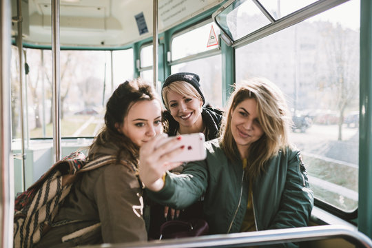 Three beautiful young women sitting in tram, chatting and taking selfie. Selective focus on girl in middle.