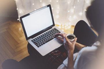 Hipster girl using computer and drink cup coffee in home atmosphere, person holding laptop on background glow bokeh Christmas illumination, hands texting on relax glitter decoration, mockup templates